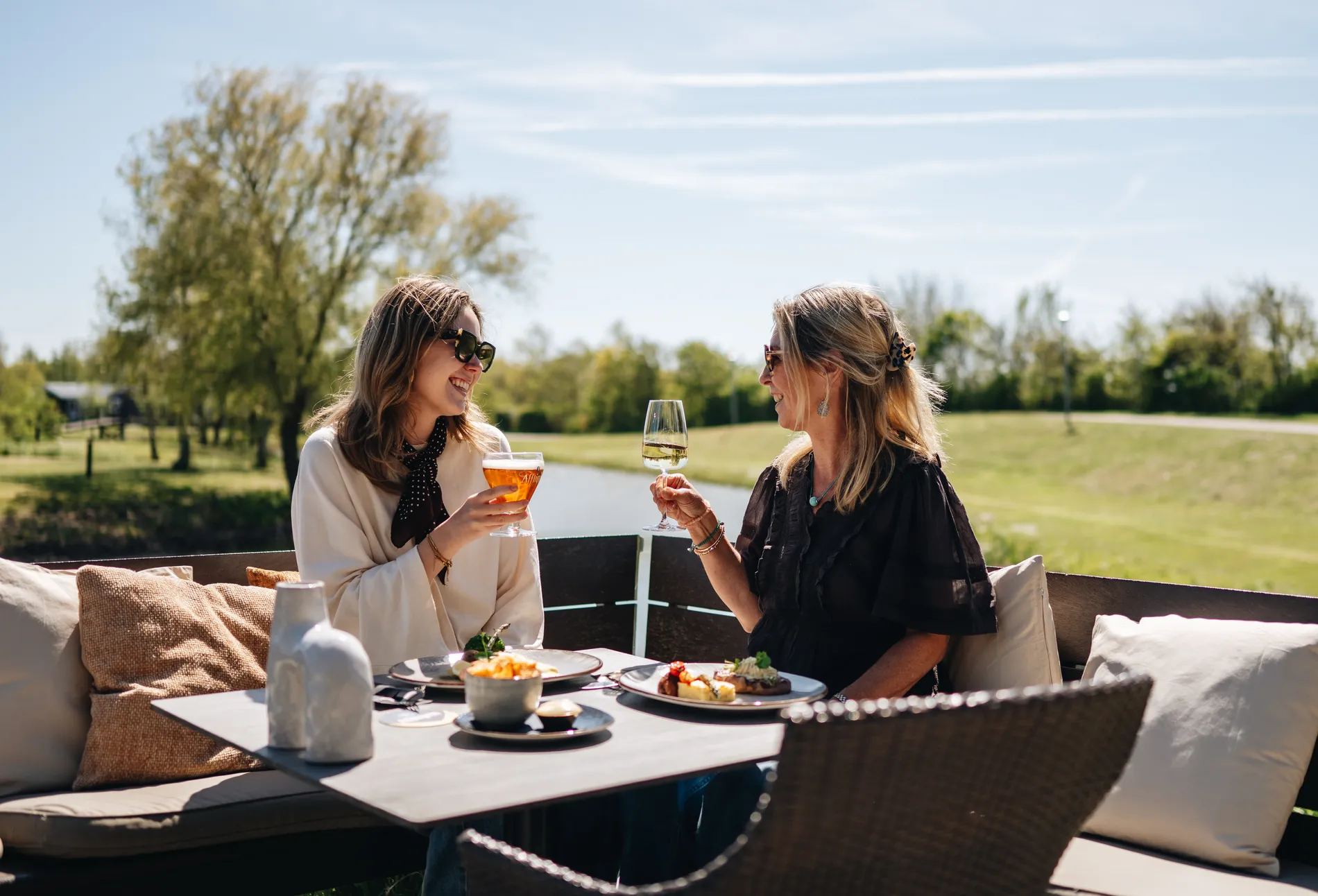 Twee lachende vrouwen proosten met een glas bier en witte wijn tijdens een lunch op het terras aan het water van het resort.